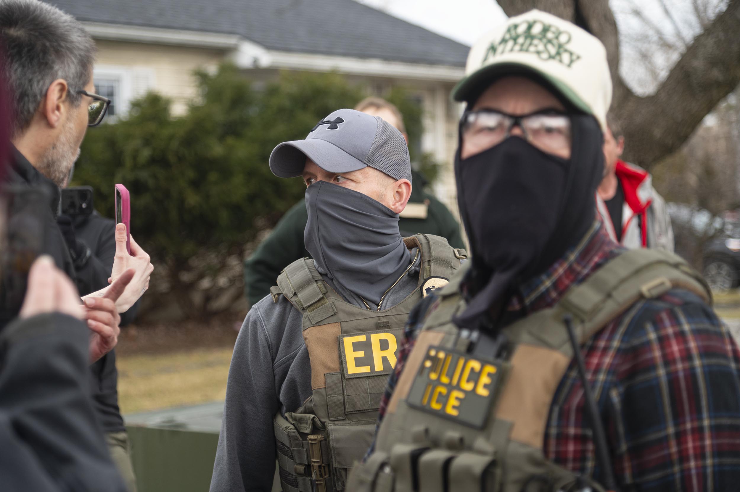Two people wearing tactical vests labeled "POLICE ICE" and "ERS" stand outdoors, one wearing a mask and hat, as another person gestures with a smartphone nearby.