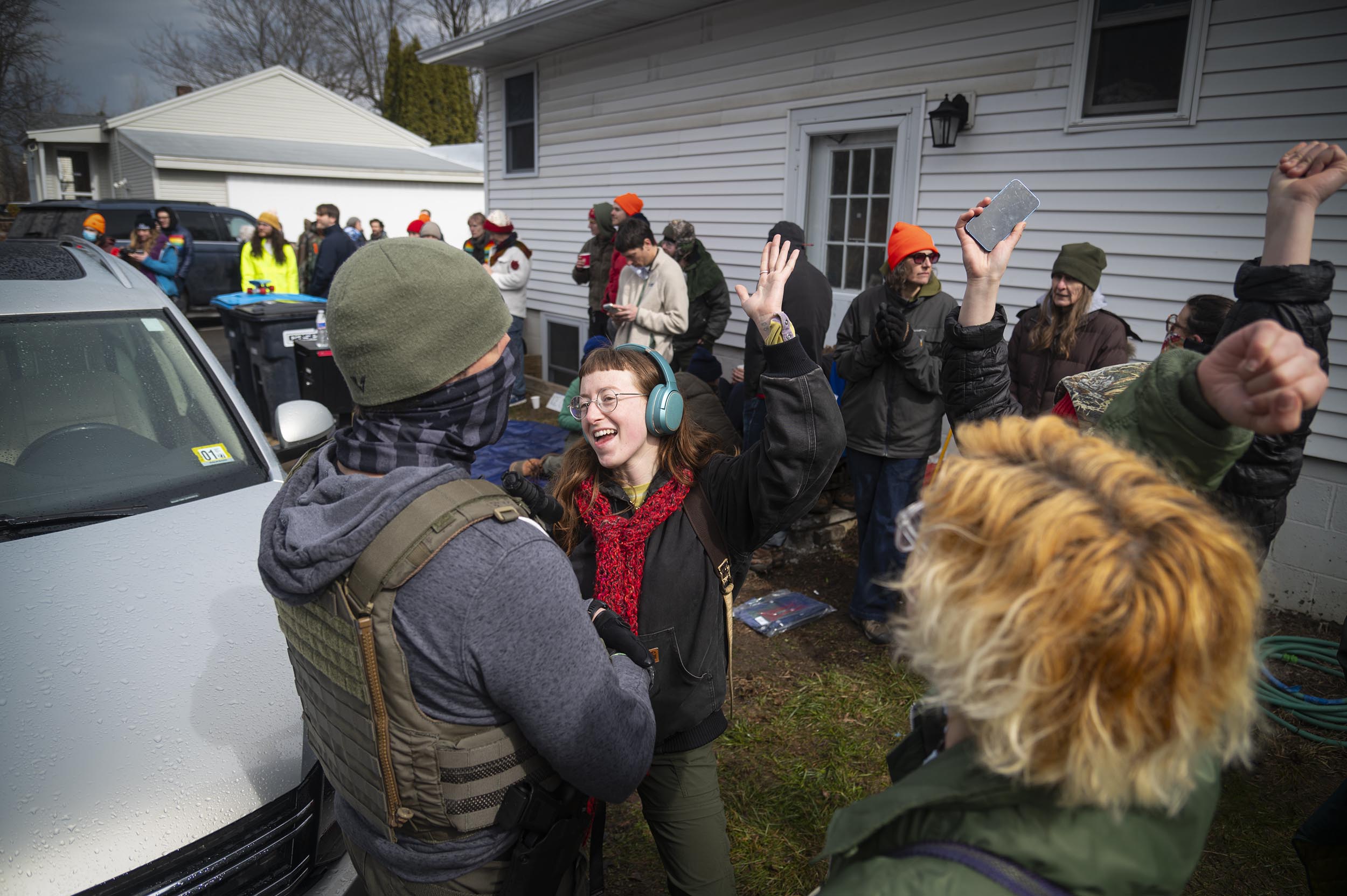 A group of people outdoors near a white house, some raising their arms and wearing headphones or hats, while two people in the foreground interact and smile.