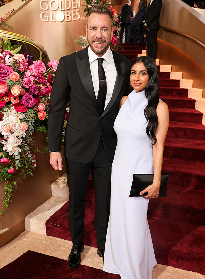Dax Shepard posing in a black suit with a woman in a white gown at a red carpet event with floral decorations.