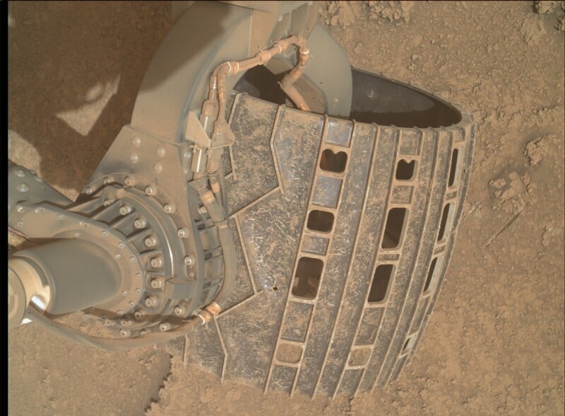 Close-up of a rover wheel on Mars, showing its metal surface covered with dust and dirt. The tread and grooves are visible, as well as mechanical parts and bolts connecting the wheel to the rover.