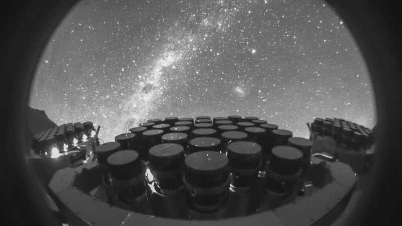 A wide-angle view of a telescope array under a star-filled night sky, with the Milky Way and clouds of stars visible above the circular arrangement of telescope lenses in the foreground.