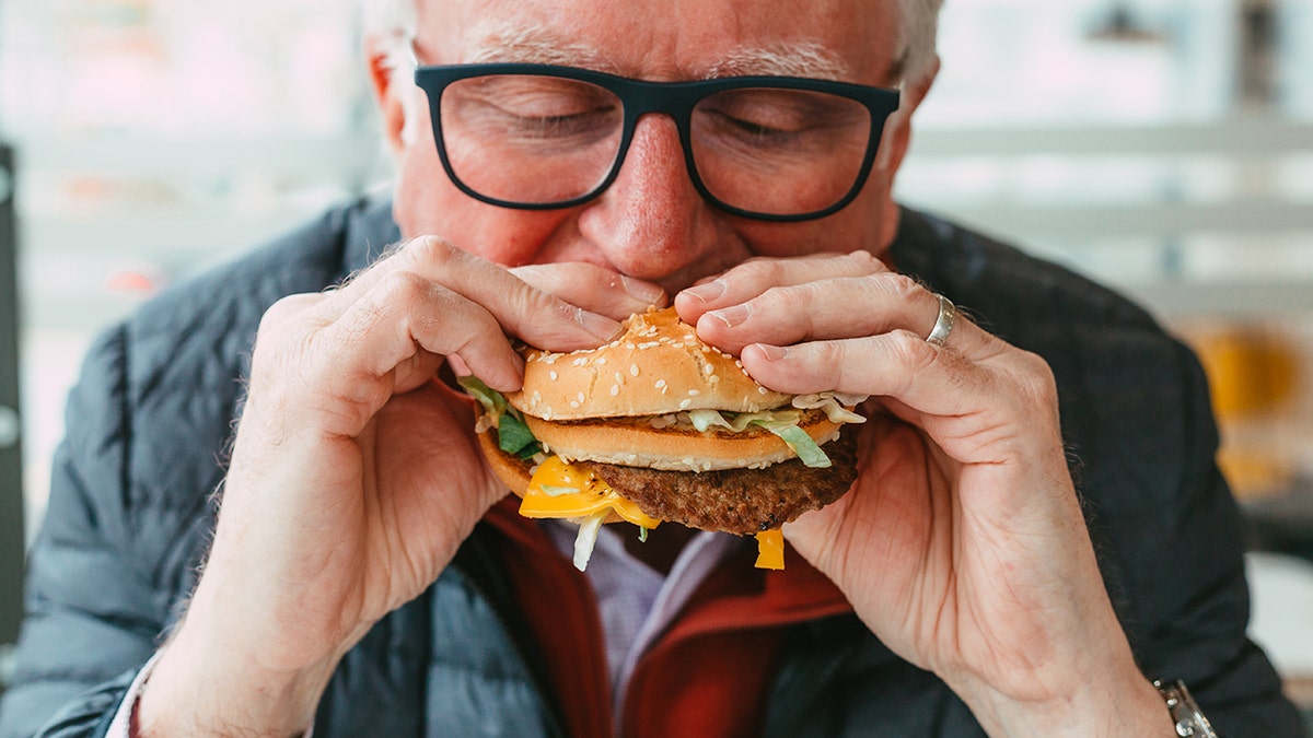 Older man with glasses eating a cheeseburger.