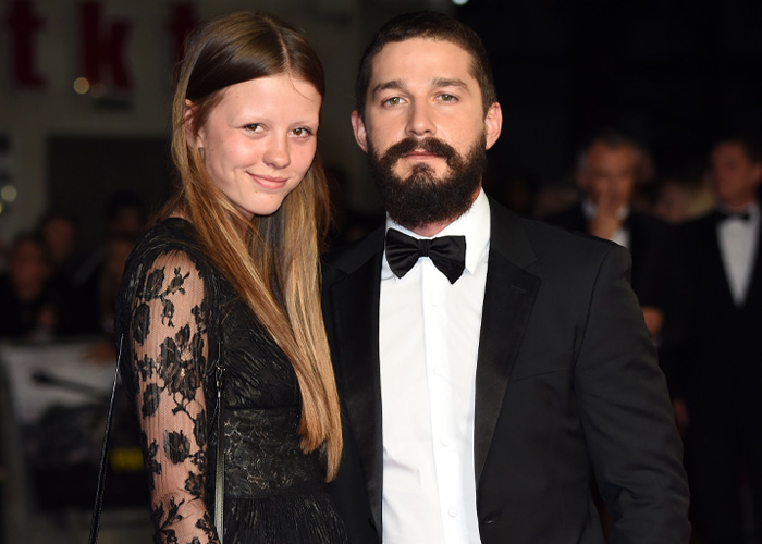 Shia LaBeouf wearing a black tuxedo and bow tie at an event, posing with a woman in a black lace dress.