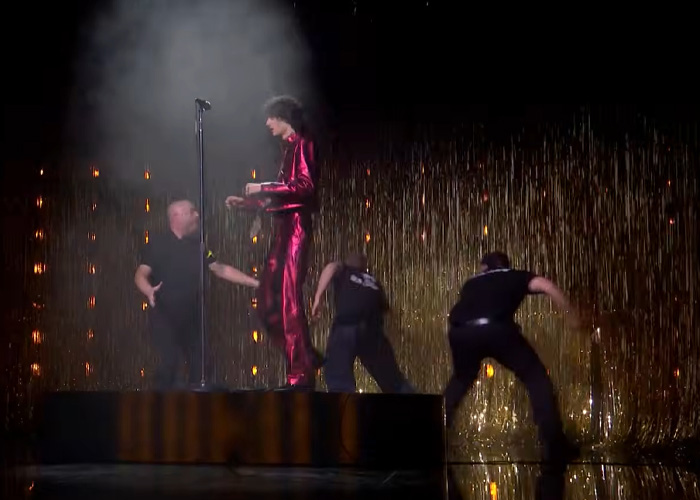 Singer in red outfit being pushed off stage during chaotic Brit Awards performance with security and gold curtain backdrop.