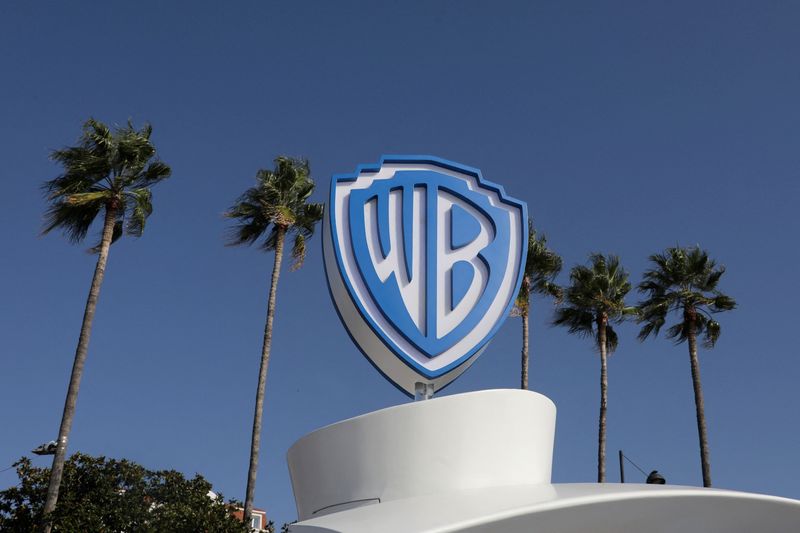 The Warner Bros. logo, a blue and white shield, stands against a clear blue sky with palm trees in the background.