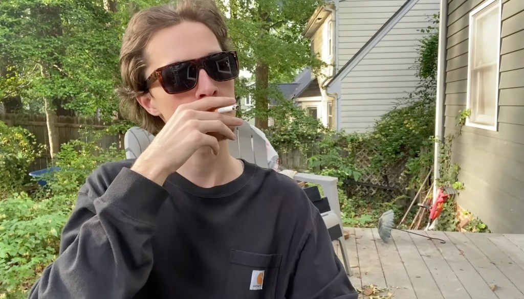 A man in a black shirt and sunglasses smokes outside. 