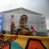 Olga Urbina holds her 9-month-old son, Ares Webster, in a front-pack baby carrier at a protest in front of the Supreme Court building. Ares holds a small American flag. Another protester holds a sign that says, "American Born Children Are American Children."