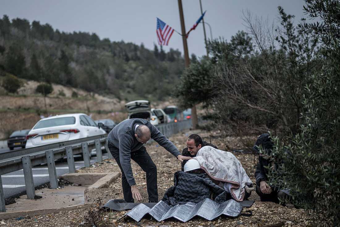 A man holds in place a protective helmet on the head of a child as other motorists take shelter from an incoming missile attack in a ditch on the side of the highway in Latrun on April 1, 2026.