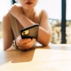 A young woman sits at a sunlit table, holding a smartphone with a soft focus background. The scene conveys casual connectivity, focus on the device, and a relaxed moment of everyday life.