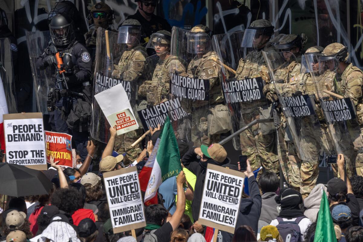 People holding signs face a row of uniformed guards in helmets, holding shields with the words California National Guard