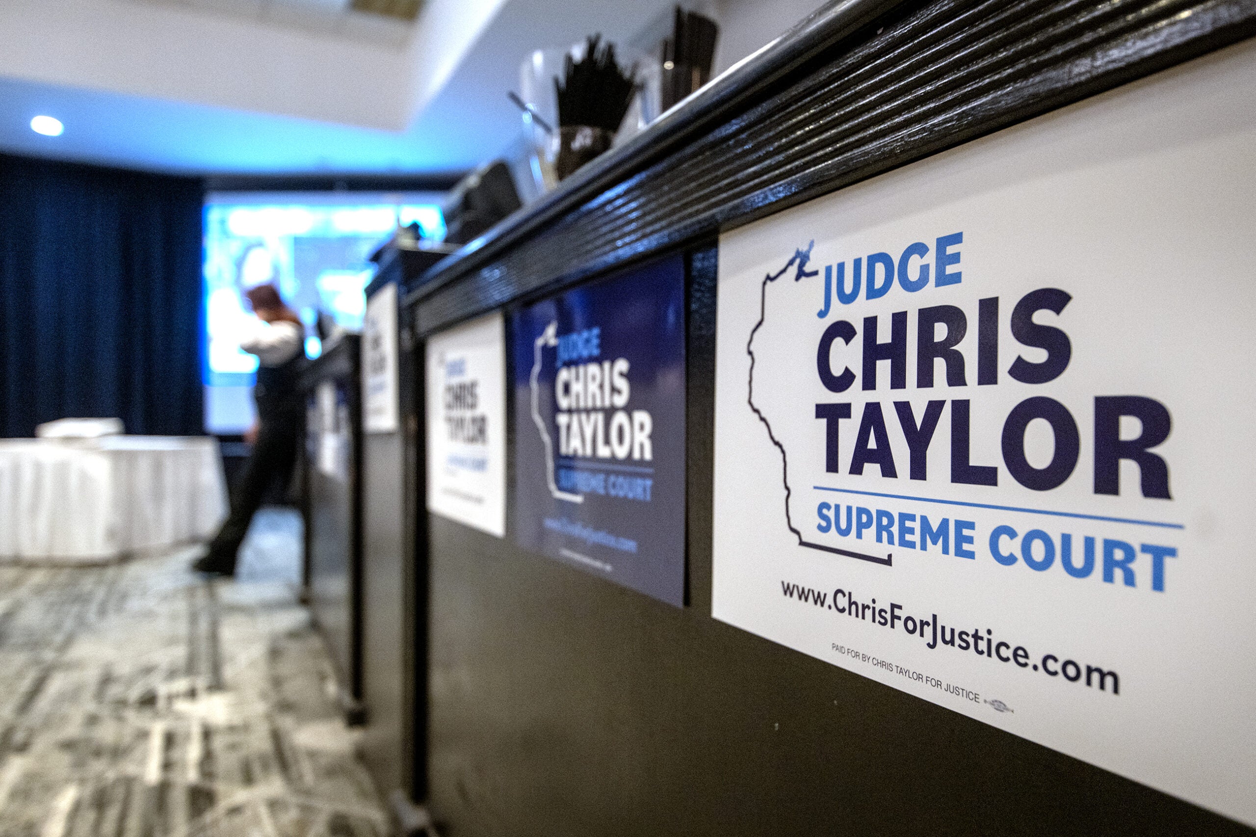 A campaign sign for Judge Chris Taylor for Supreme Court is posted on a podium at an indoor event, with a person and presentation screen visible in the background.