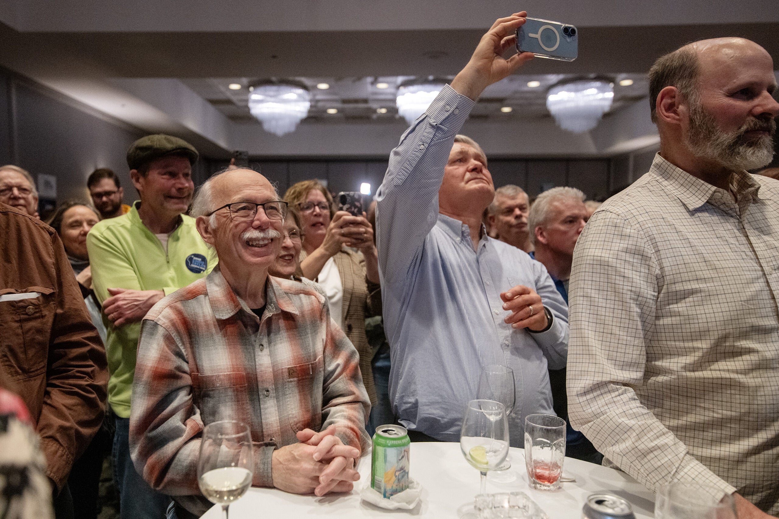 A group of people stand closely together in a room, some smiling and some photographing or recording with phones; drinks and glasses are on a table in the foreground.