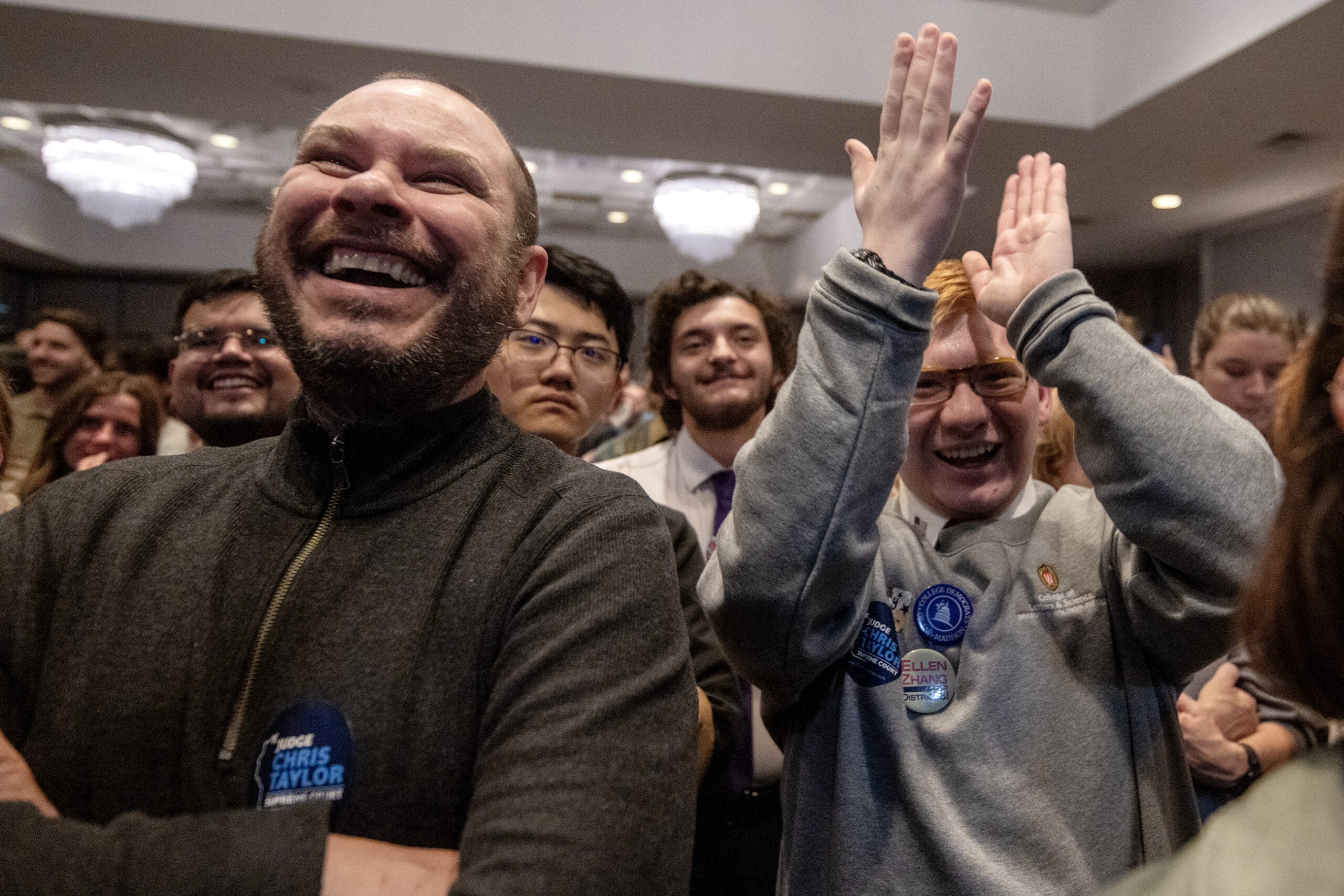A group of people indoors smile and cheer, some wearing campaign buttons and stickers, suggesting a celebratory or political event.