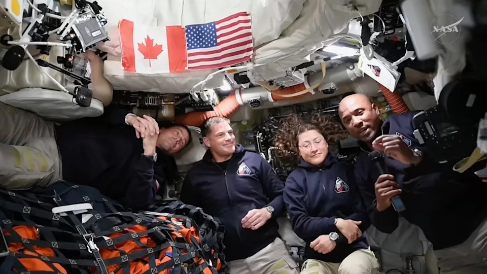 three male and one female astronaut, all wearing blue jackets and khaki pants, smile for the camera inside their space capsule in space