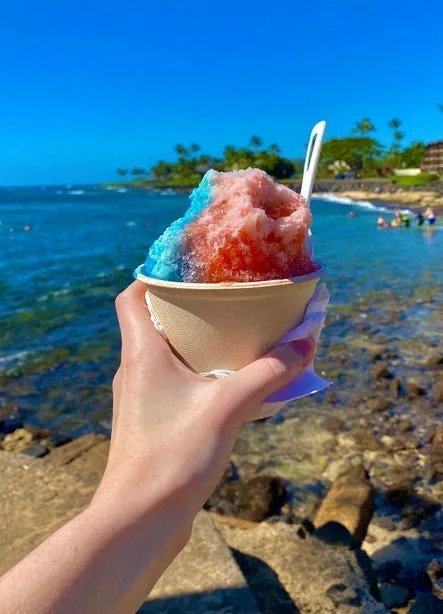 Person holding a cup of shaved ice with red and blue sections at a beach, with ocean and people in the background