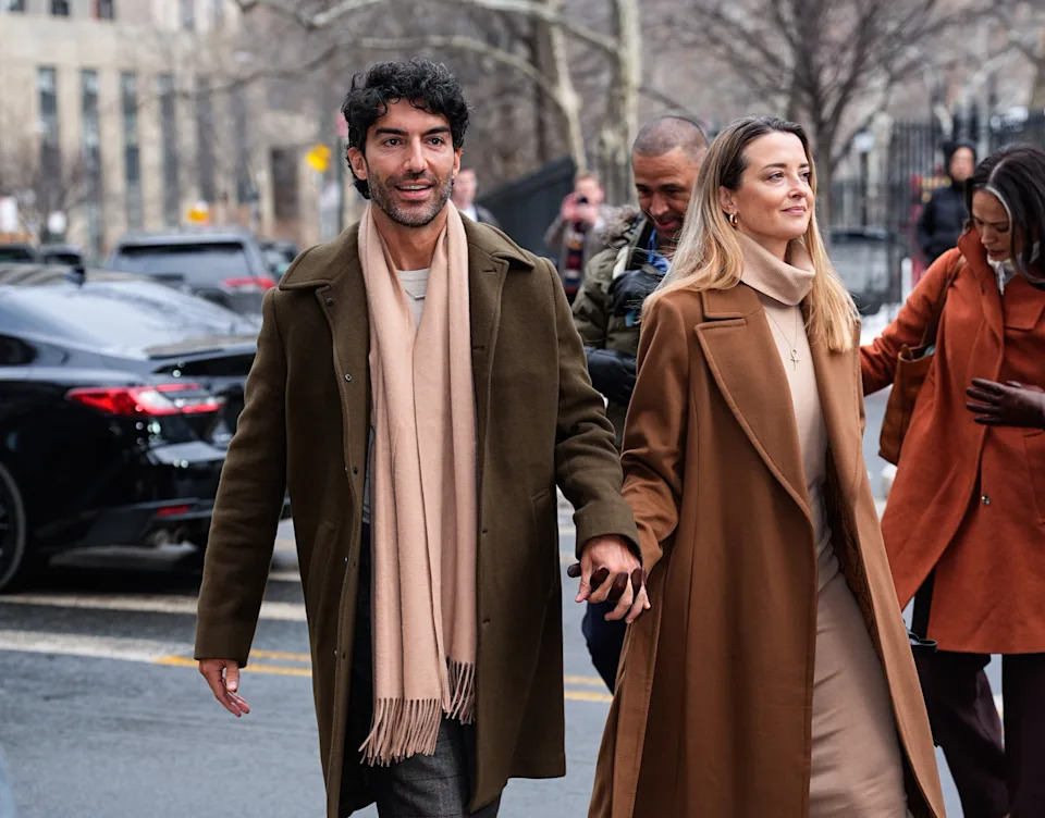 A couple walks hand in hand in stylish coats on a city street, smiling towards the camera