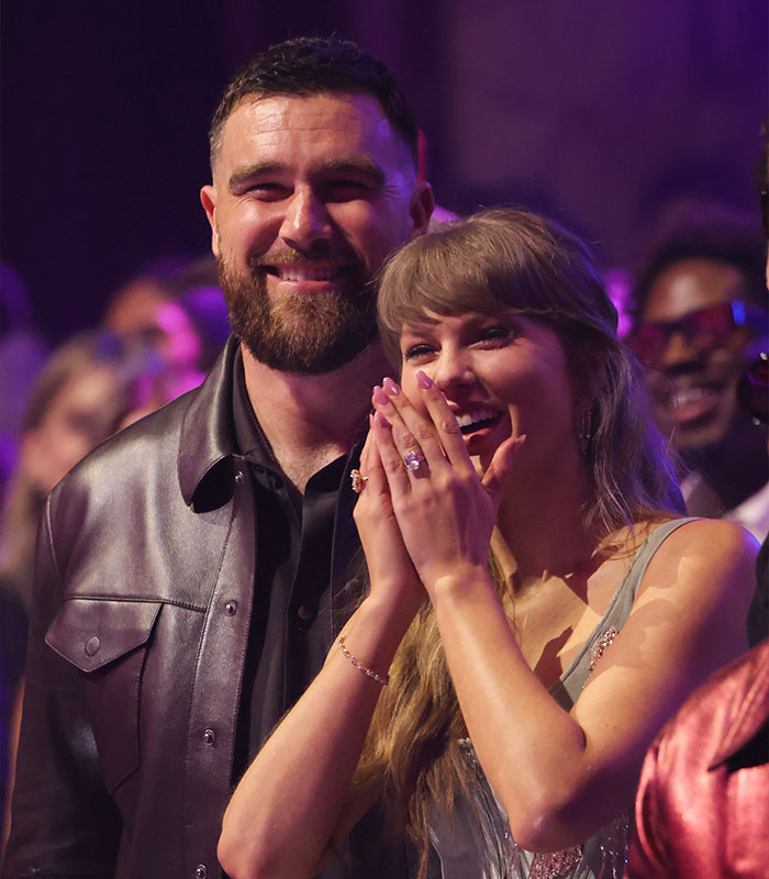 Travis Kelce smiling beside Taylor Swift at an event, with fans and purple lighting in the background.