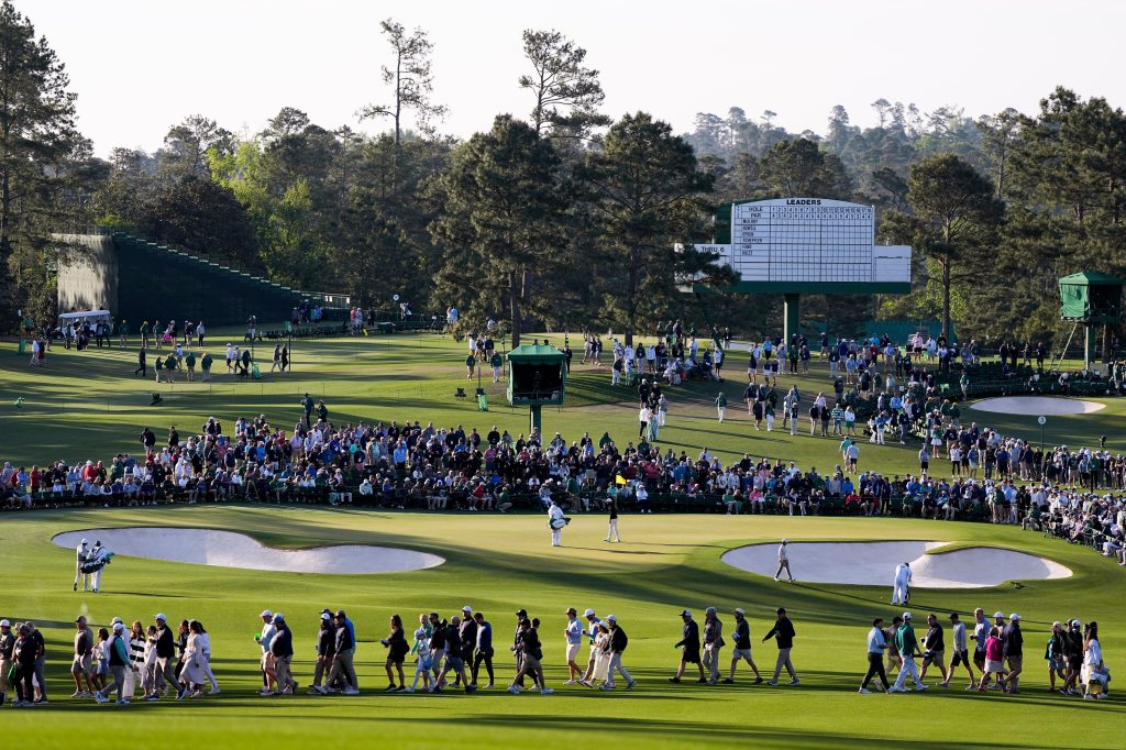 A large crowd watches golfers on a course at the Masters Golf Tournament, with a scoreboard displaying "LEADERS" in the background.