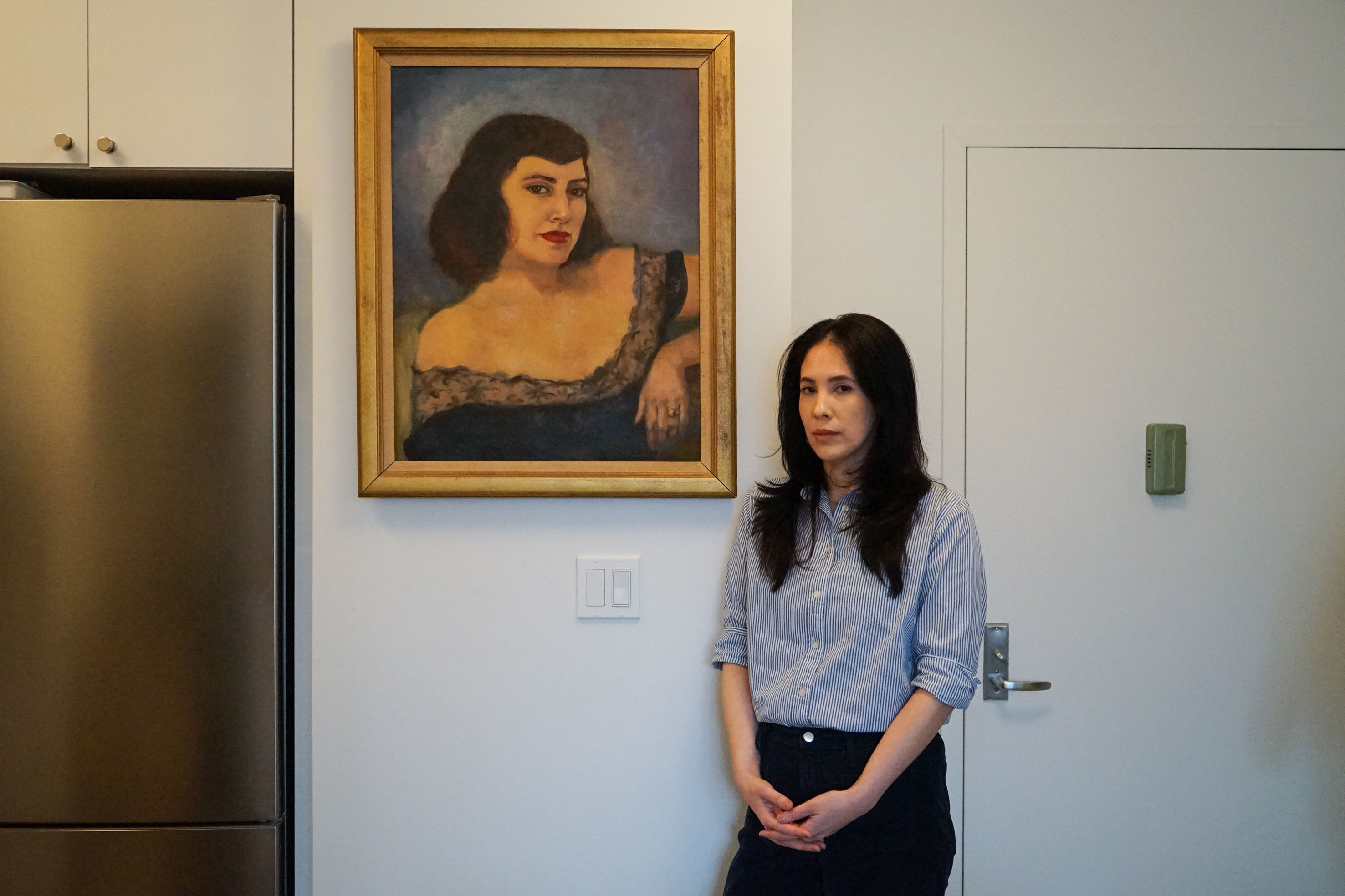 A woman stands beside an oil painting of her grandmother as a young woman. They resemble one another.