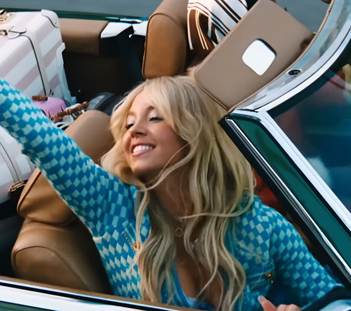 Sydney Sweeney smiling and reaching out while sitting in a convertible car, showcasing her look in the new Euphoria season.