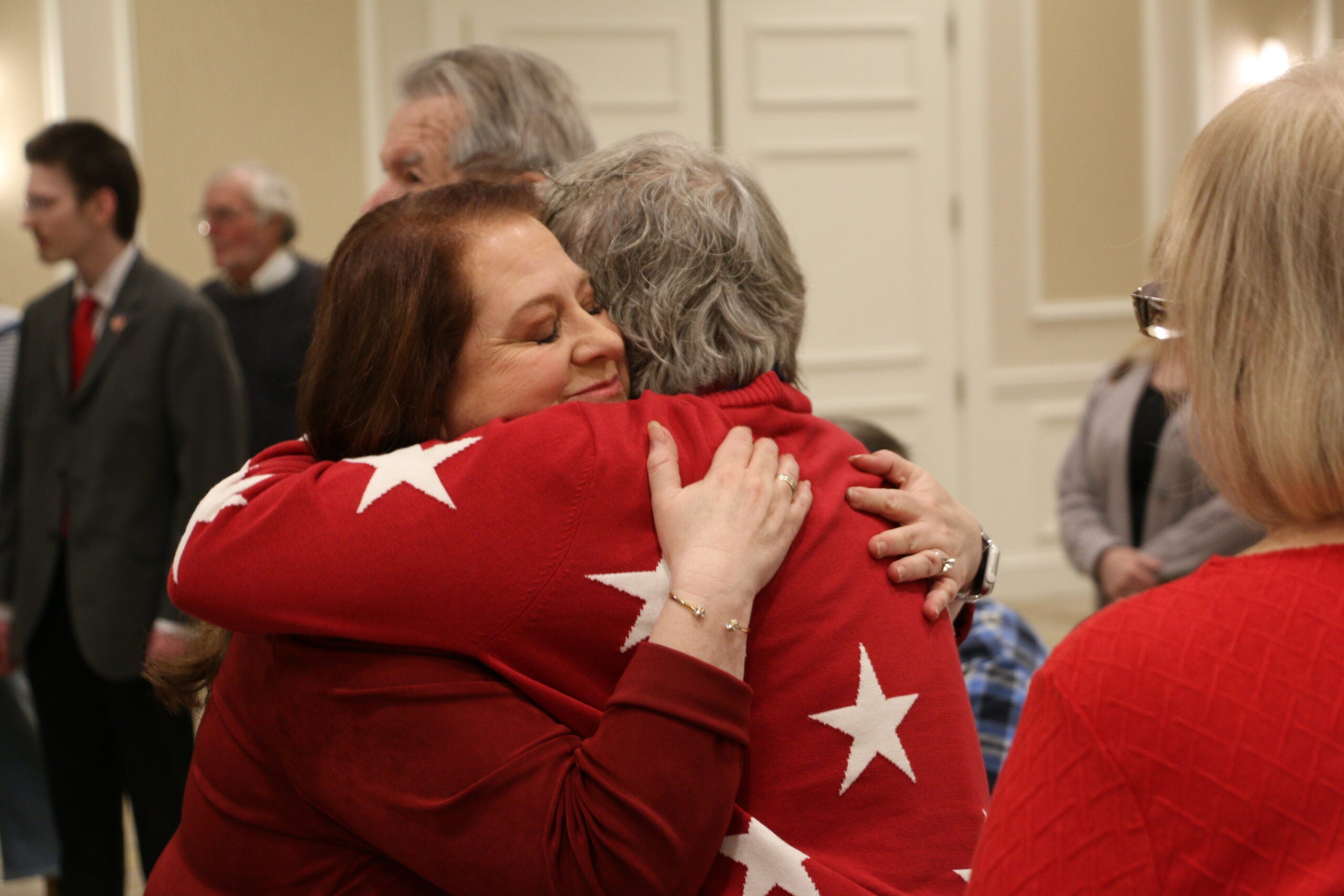 Two women wearing red sweaters with white stars hug each other in a crowded indoor setting, while other people stand and talk in the background.