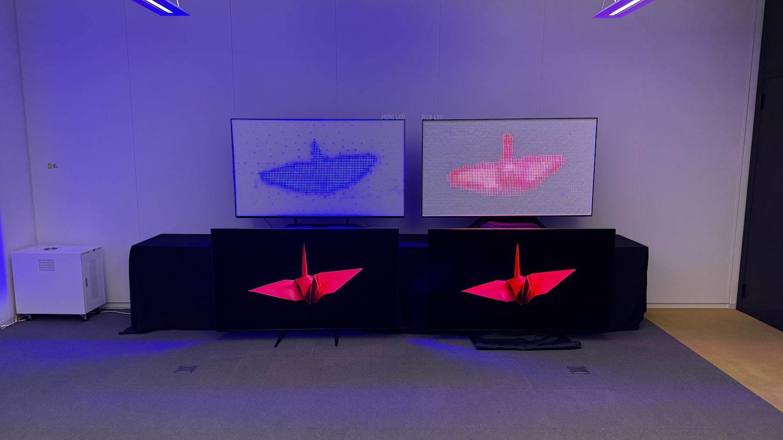 Two TVs arranged next to one another on the floor, with the exposed backlights of each on a table above. On both of the assembled TVs, an image of an origami bird is displayed. On the left-side backlight, a vague blue/white image is visible. On the right-side backlight, the colours are full recreated and the shape of the bird is clear.
