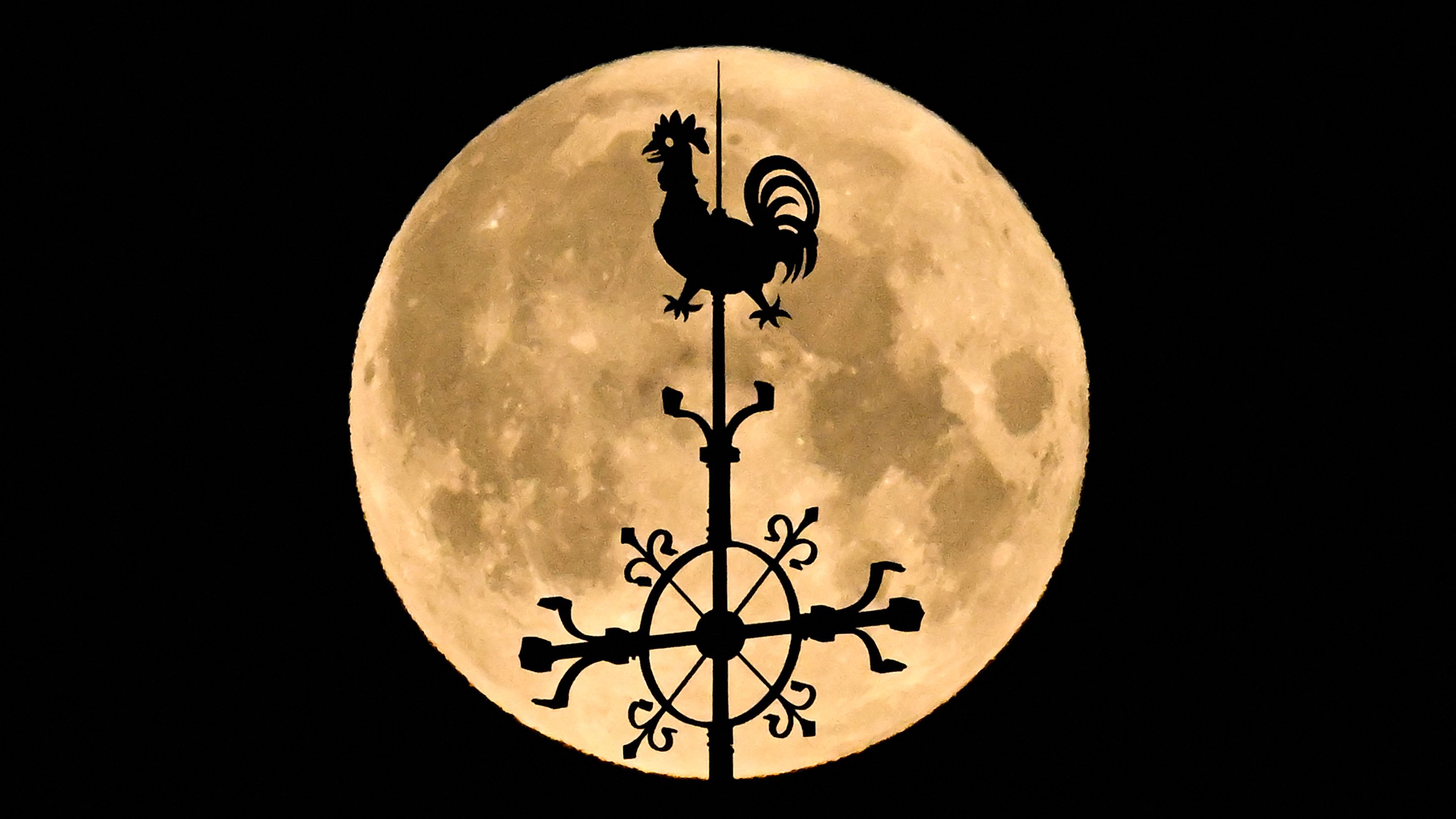 The silhouette of a weathervane is pictured In front of a yellow full moon in a black sky.