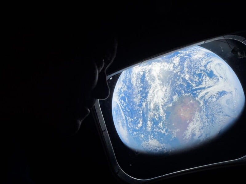 A person’s silhouette is seen looking out of a spacecraft window at Earth, which appears bright and detailed against the darkness of space, showing continents and swirling clouds.