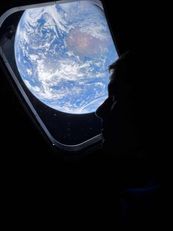 A person’s silhouette is seen looking out of a spacecraft window at the Earth, which appears large and vibrant against the darkness of space.