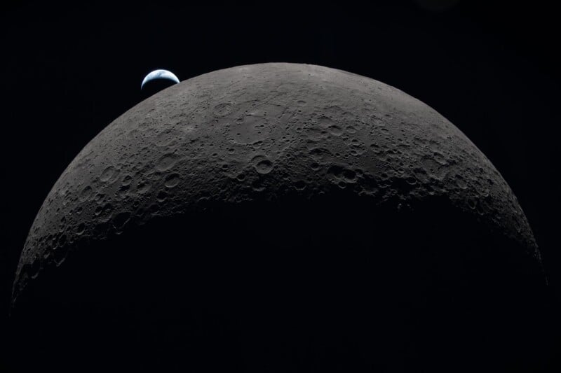 A close-up view of the moon’s cratered surface in deep shadow, with the distant Earth partially visible rising over the lunar horizon against the blackness of space.