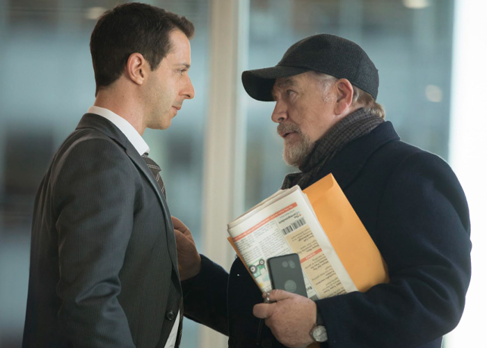 Brian C*x in a dark coat and cap, holding documents and phone, having an intense conversation with a man in a suit.