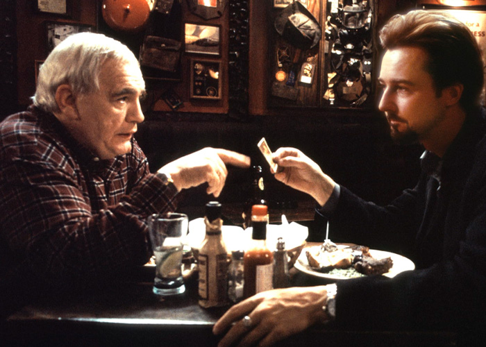 Two men having a tense conversation across a table in a dimly lit restaurant, highlighting Brian C*x unhinged backlash.