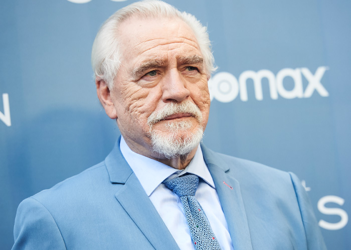 Brian C*x wearing a blue suit and tie at an event, facing backlash after a brutal dig in the entertainment industry.