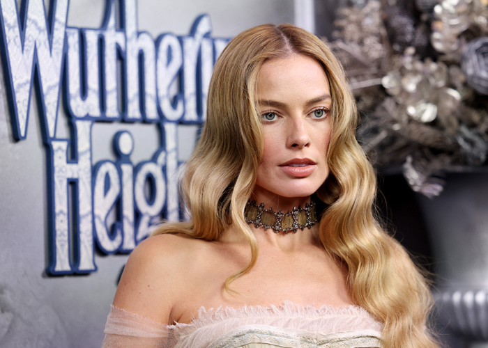 Margot Robbie posing at a red carpet event with wavy blonde hair, linked to Brian C*x backlash headlines.