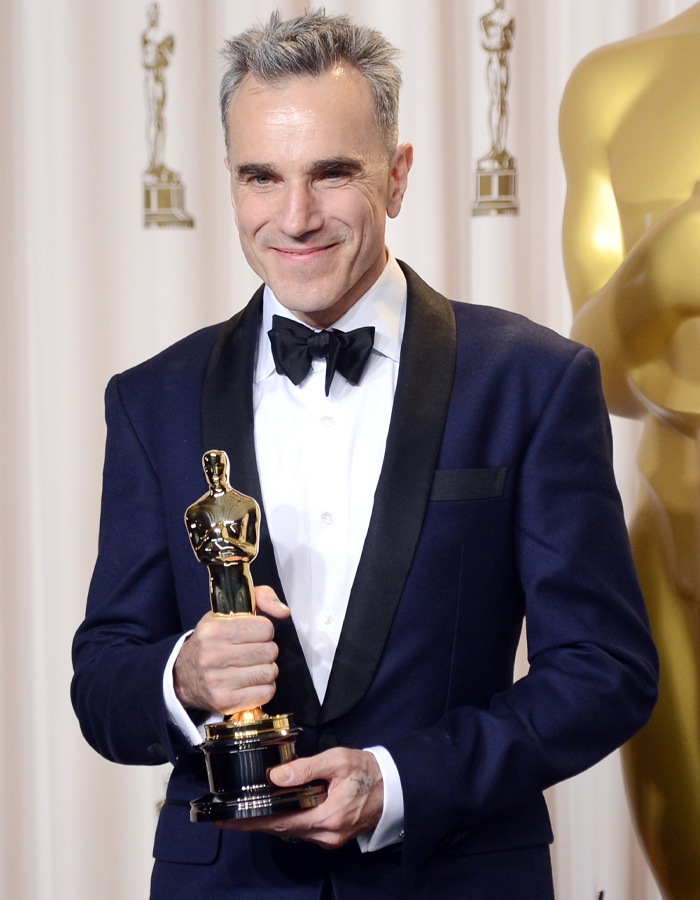 Brian C*x in a tuxedo holding an Oscar statue at an awards ceremony, facing backlash in the entertainment industry.