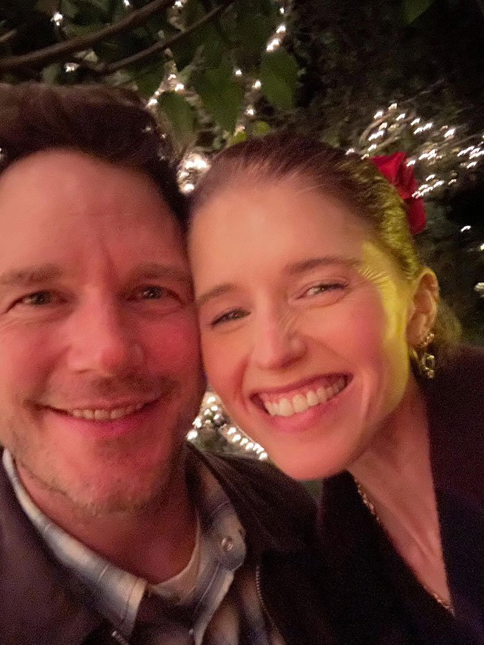 Katherine Schwarzenegger and husband Chris Pratt smiling outdoors with festive string lights in the background at night.