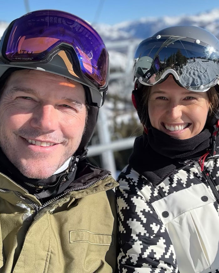 Katherine Schwarzenegger and Chris Pratt smiling outdoors in ski gear with helmets and goggles on a sunny winter day.