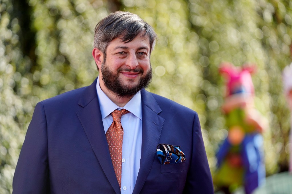 Eugene Mirman, wearing a blue suit, orange tie, and pocket square, smiles at the camera.