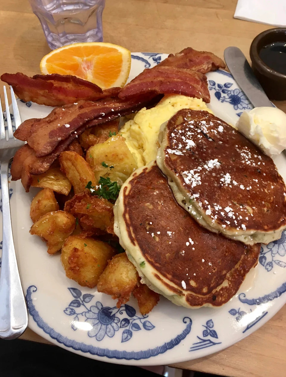 A hearty breakfast plate with pancakes, bacon, scrambled eggs, crispy potatoes, and sliced orange, served with butter on a floral plate