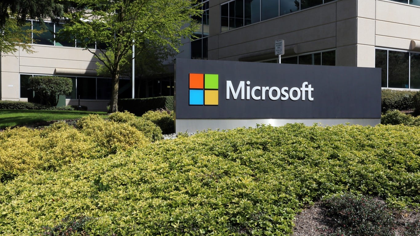 A large, dark grey sign displaying the colorful Microsoft logo and the company name in white text, set in front of multi-story beige office buildings with dark windows. The sign is surrounded by green and yellow shrubs and trees.