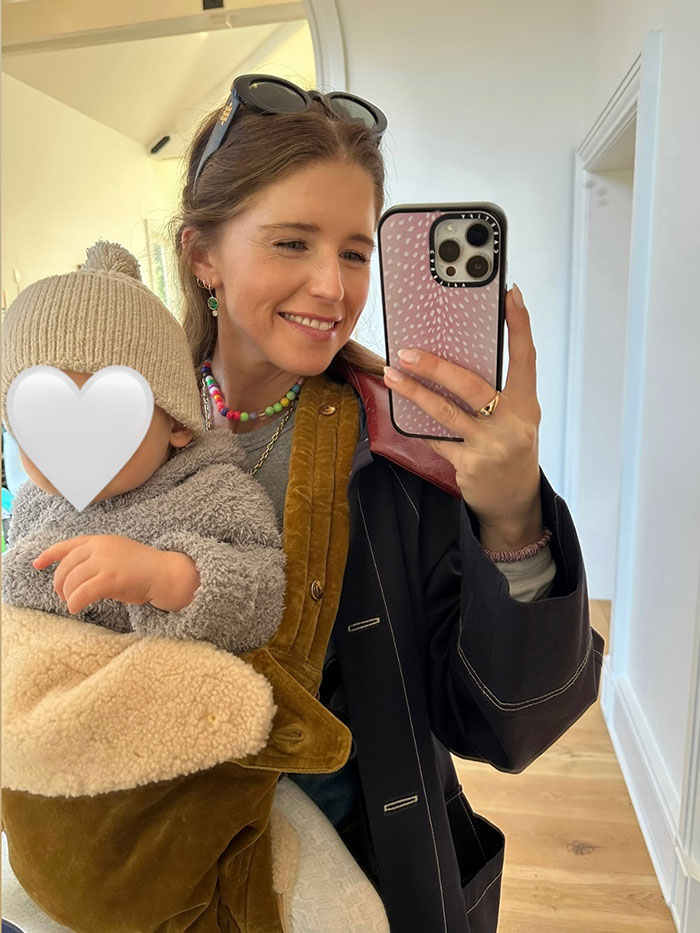 Katherine Schwarzenegger holding her child, taking a mirror selfie while wearing a colorful necklace and casual outfit indoors.