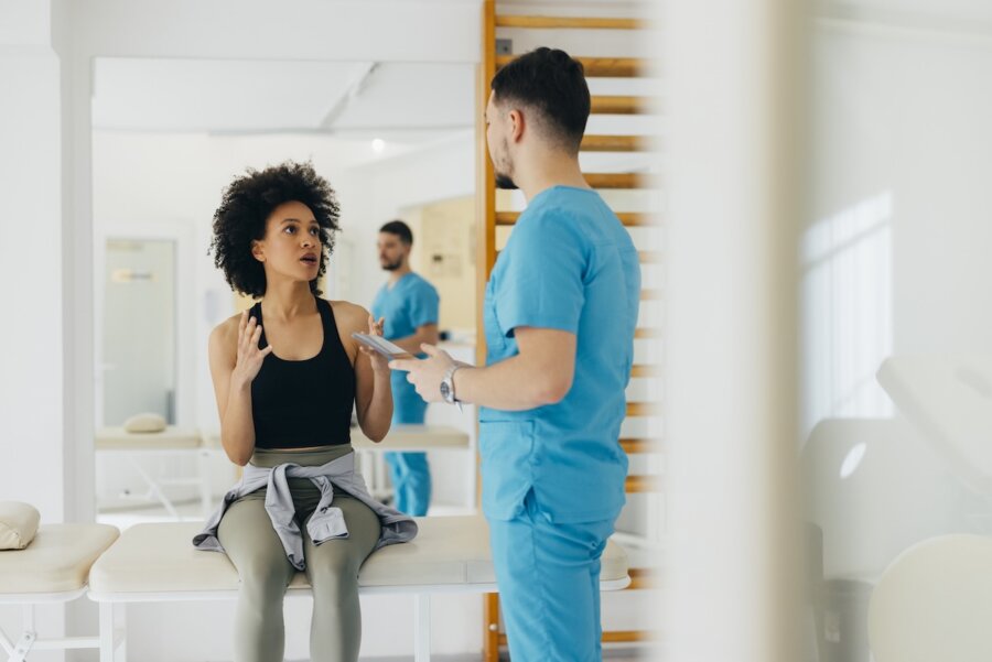 A woman sits on an exam table in a medical clinic, talking to a healthcare professional in blue scrubs who is holding a clipboard and discussing the potential benefits of NAD supplements.