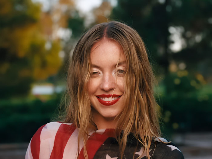 Sydney Sweeney smiling outdoors with wet hair, wearing a star-spangled outfit in a natural green background setting.