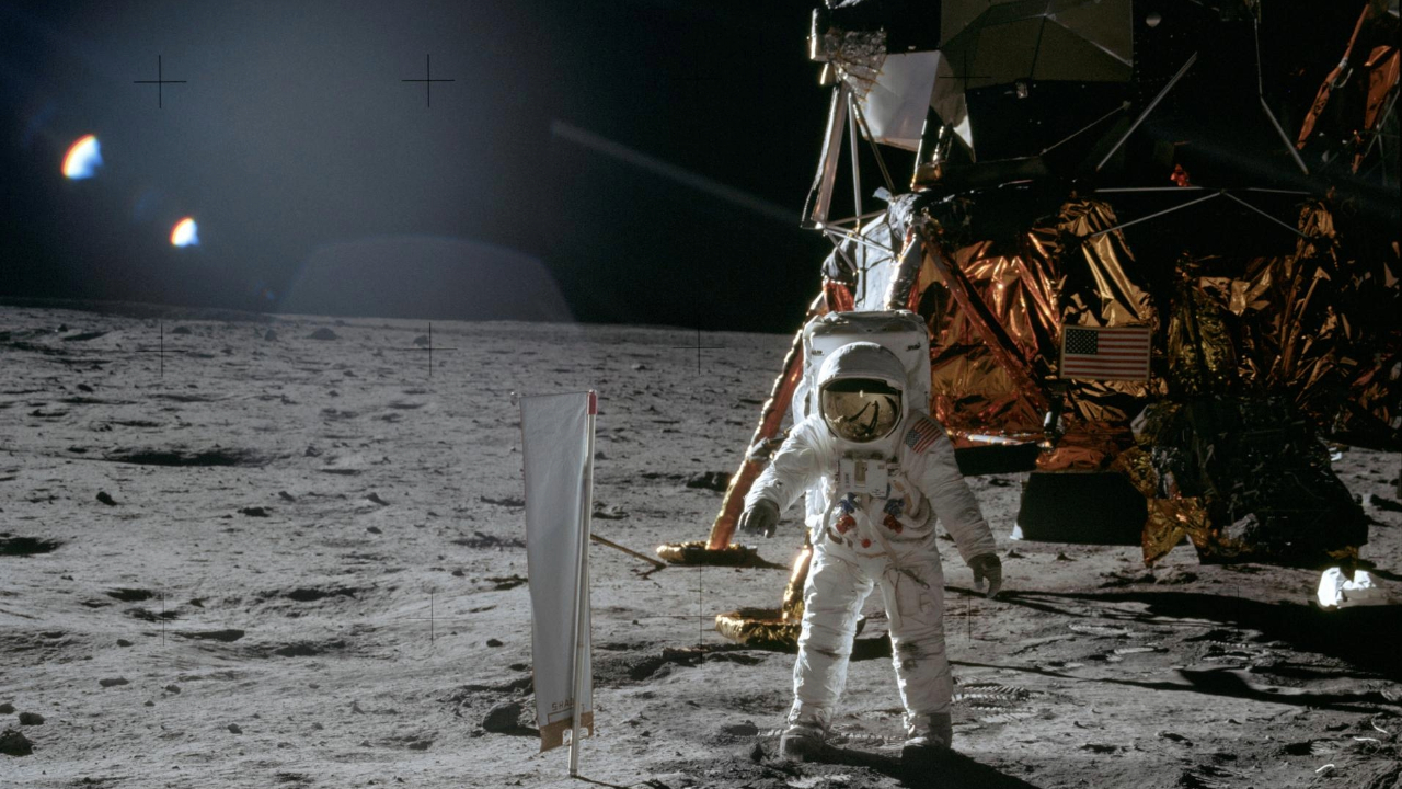 An astronaut wearing a white space suit stands next to a flag on the moon with a large golden lunar lander behind him